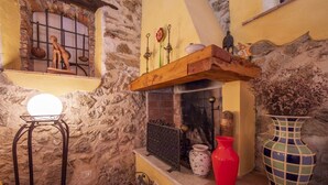 Un interior rústic amb una paret de pedra, una estanteria de fusta amb objectes decoratius, una llar de foc i un vas vermell.