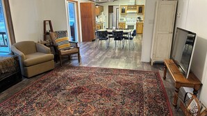 Una sala de estar con un tapete grande de estampado, un sillón color beige, una mesita de madera con un televisor, y un comedor al fondo.