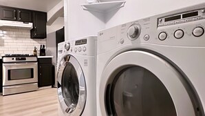 Un moderno cuarto de lavandería con dos lavadoras de carga frontal, un estante pequeño encima de las máquinas y una zona de cocina al fondo.