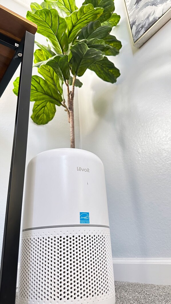 Un purificador de aire Levoit blanco con etiqueta ENERGY STAR azul, colocado sobre un piso alfombrado junto a una planta y un cuadro en la pared.