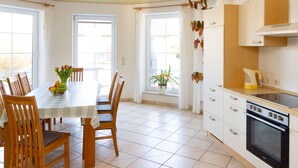 Une salle à manger lumineuse, avec une table et des chaises, une cuisine équipée d’une cuisinière et de placards, et de grandes fenêtres avec des rideaux.