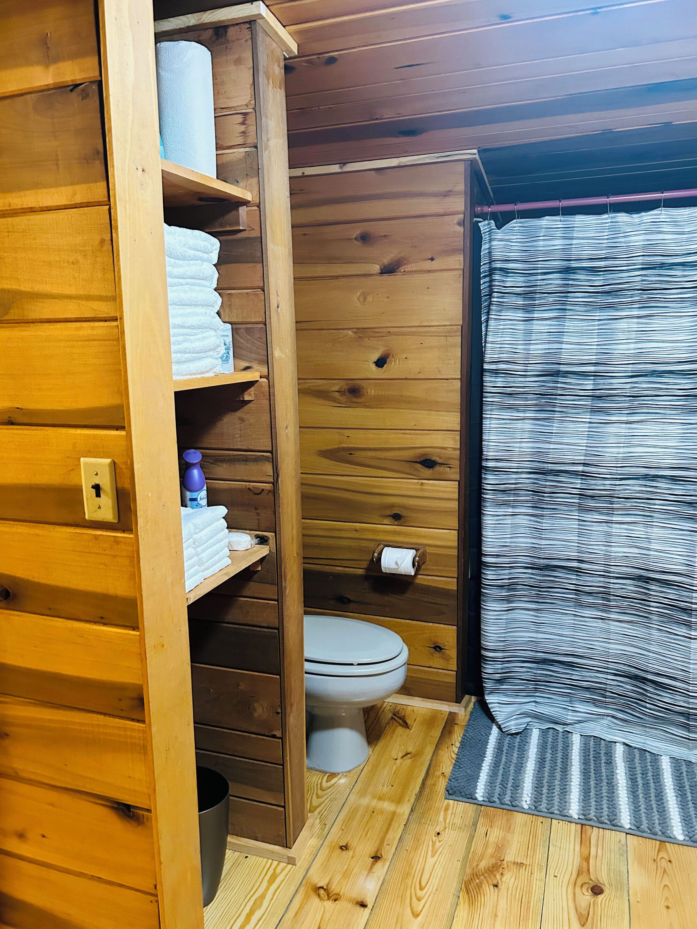 A wooden cabin bathroom with a toilet, shelves with towels, and a shower curtain.