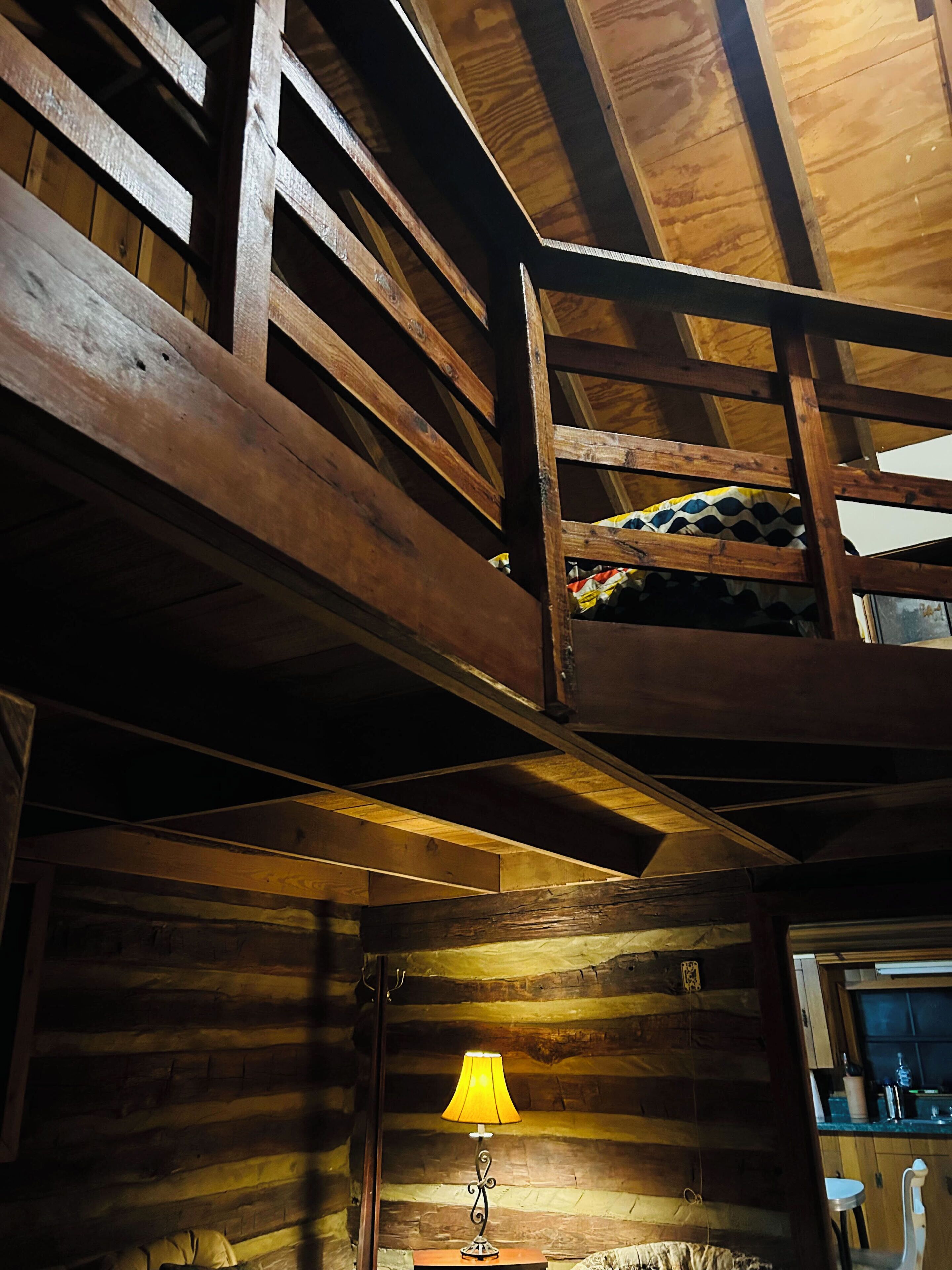 A rustic cabin interior with a loft bed, a wooden ceiling, and a lamp on a table.