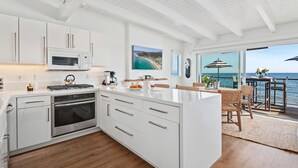 A modern kitchen with white cabinetry, stainless steel appliances, and a large island. There is a dining area with wicker furniture and a view of the sea. - Exclusive Luxury Beachfront Home With Two Separate Units (Santa Barbara)