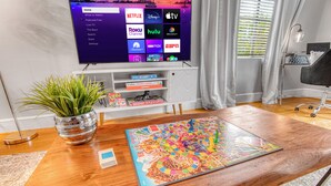 Un salon moderne doté d’un téléviseur à écran plat affichant l’interface d’un service de diffusion en continu, d’une table basse sur laquelle est posé un jeu de société, d’une plante en pot et d’une bibliothèque.