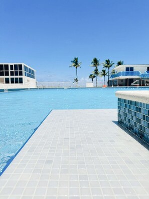 A swimming pool with a tiled deck, palm trees, and buildings in the background.