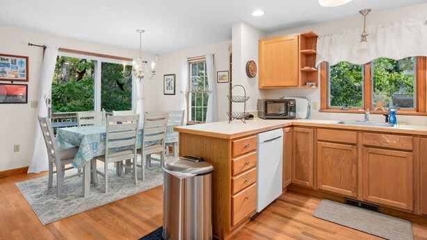A compact kitchen with wooden cabinets, a microwave, and a sink. There is a dining area with a table and chairs, and a trash can is visible. - Woods Edge: Walk to Red River Beach, Pet Friendly (Harwich Port)