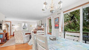 A spacious living room with a large dining table, multiple chairs, a fireplace, and a view of greenery through large windows. - Woods Edge: Walk to Red River Beach, Pet Friendly (Harwich Port)