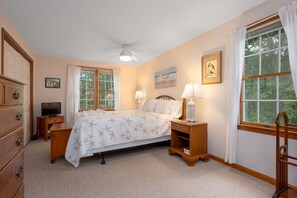 A bedroom with a bed, two bedside tables with lamps, a dresser, a television, and two windows with curtains.