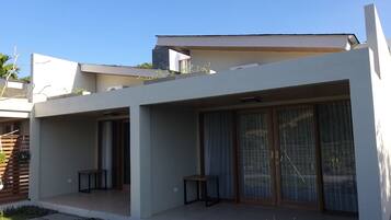 A modern two-story house with a flat roof, large sliding glass doors, and a small patio area with a table and chairs.