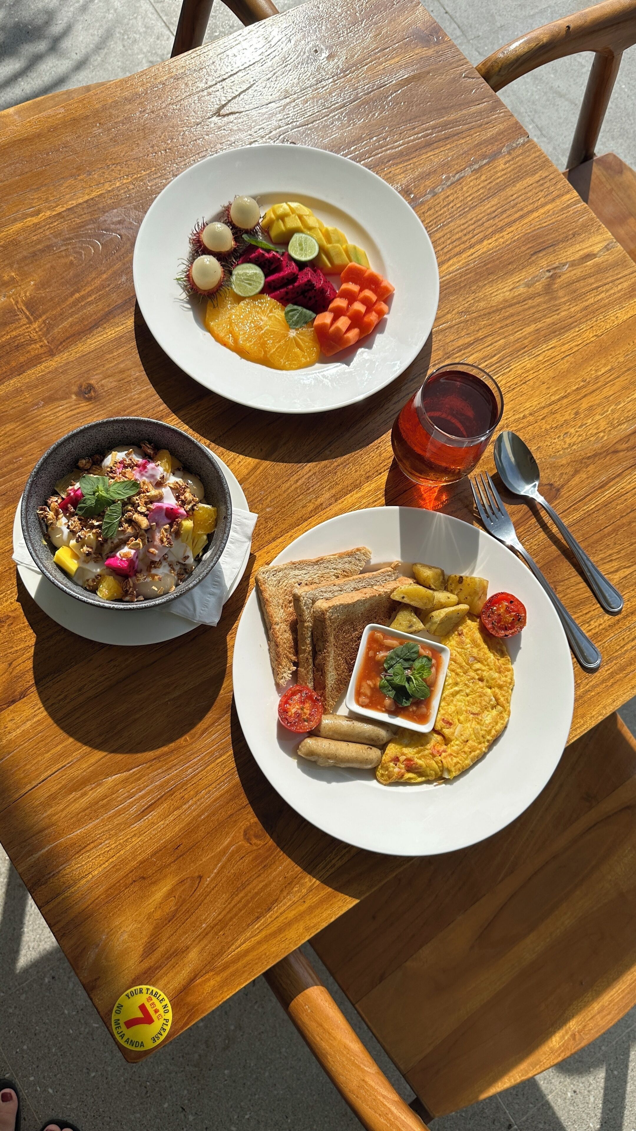 A wooden table with two plates of food, a bowl of granola, and a glass of tea.