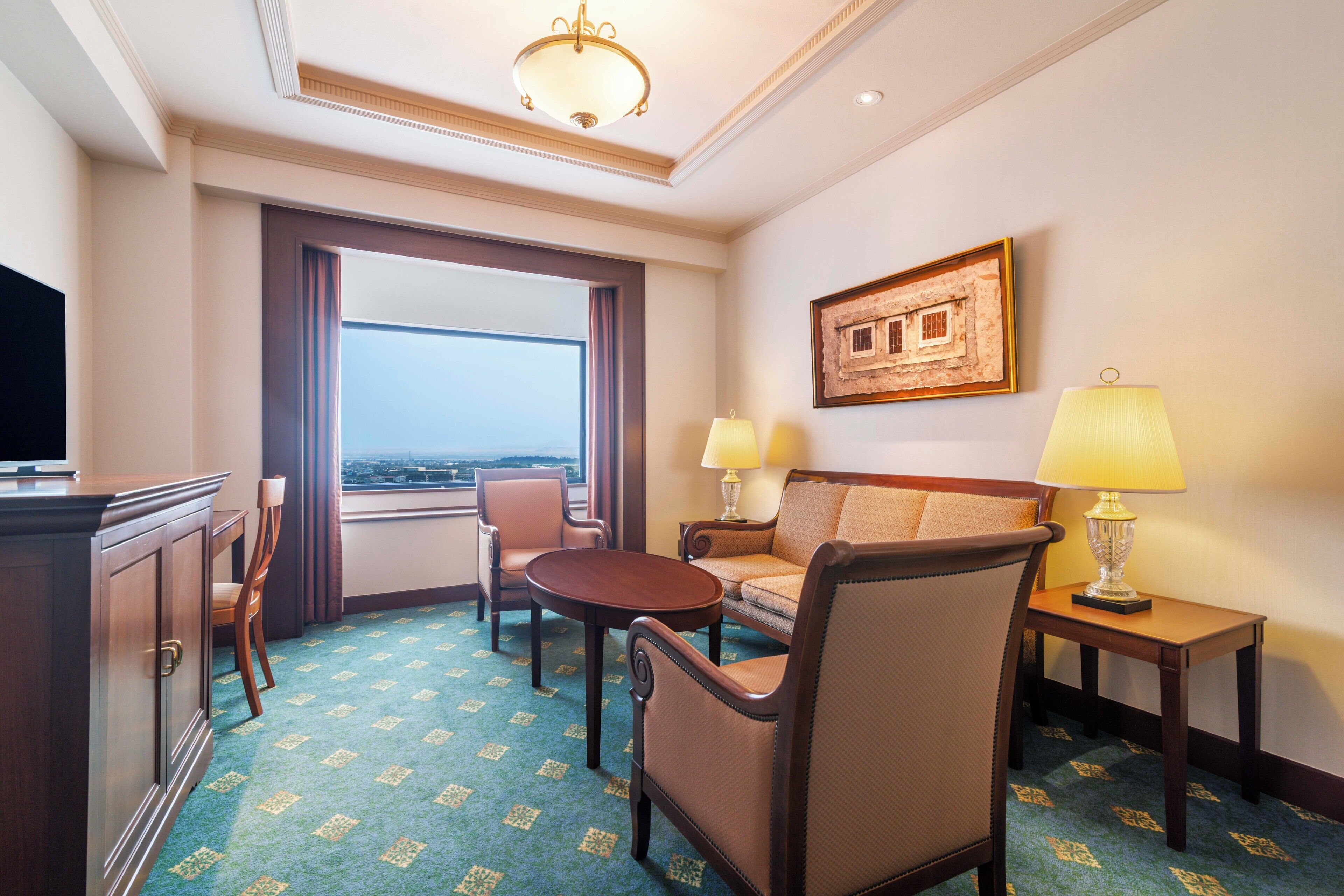 A hotel room with a large window, a sofa, a chair, a small table, and a lamp.