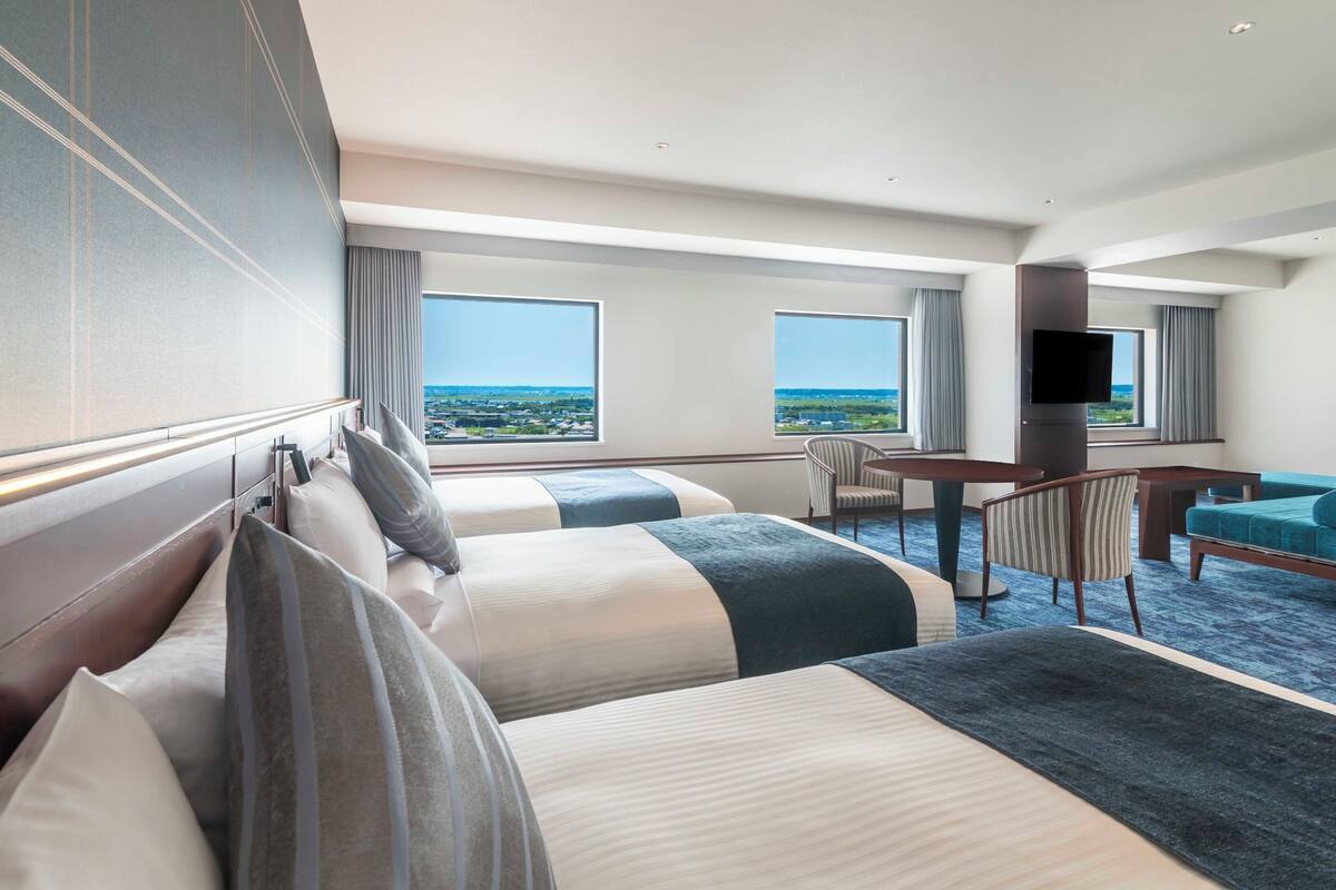 A hotel room with two beds, a desk, a chair, and large windows offering a view of the cityscape.
