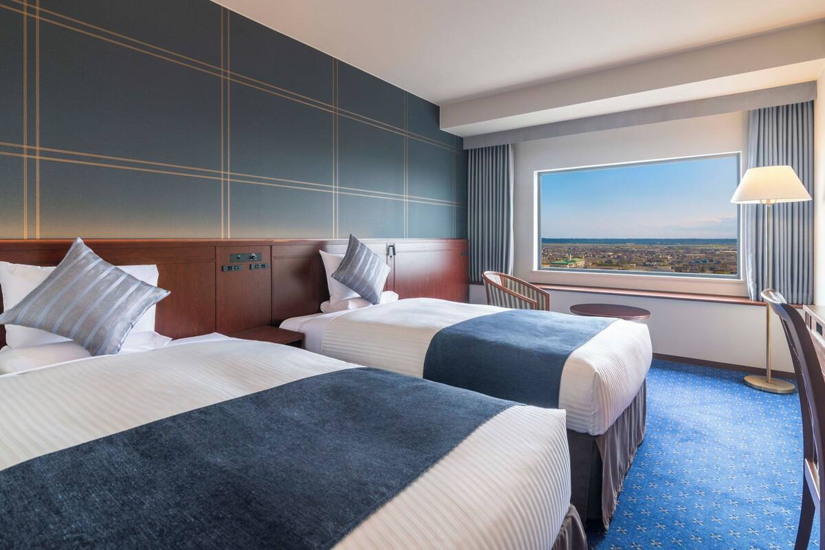 A hotel room with two beds, a desk, and a large window offering a city view.