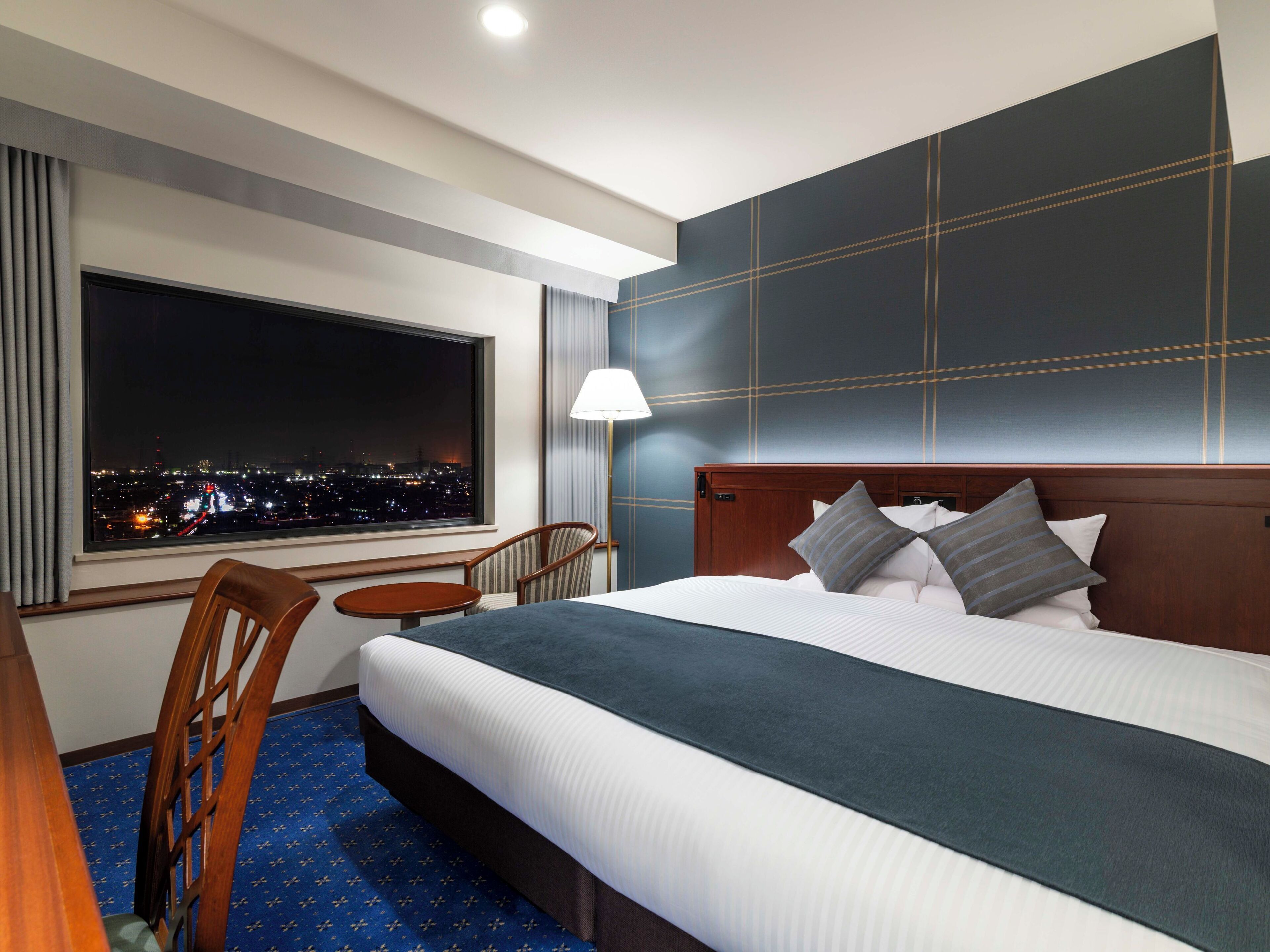 A hotel room with a large bed, a chair, a small table, and a view of the city at night.