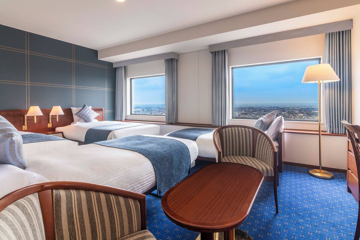 A hotel room with two beds, a desk, and a chair. There are large windows with a city view.