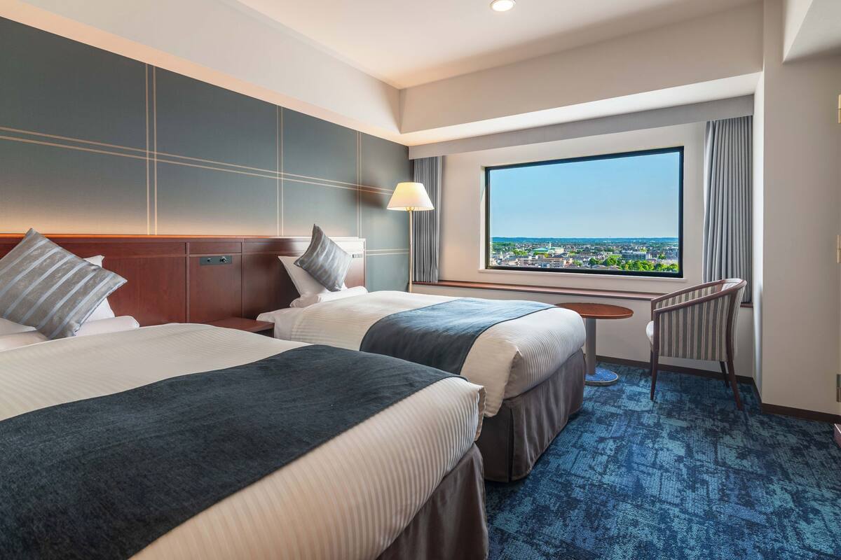A hotel room with two beds, a view of the city, a desk, and a chair.
