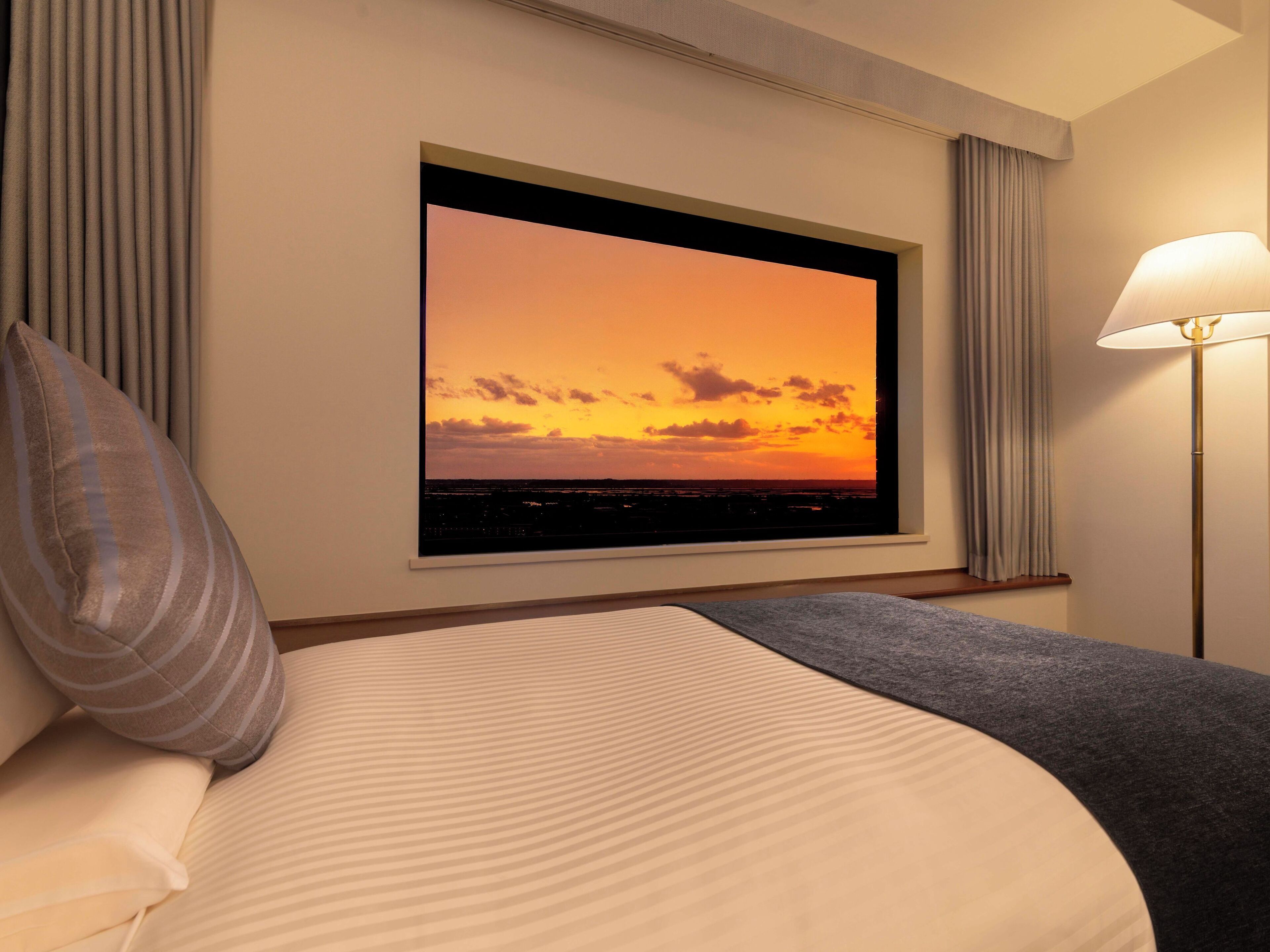 Habitación de hotel con un ventanal que muestra una vista del atardecer, una cama con una colcha gris y una lámpara de pie.