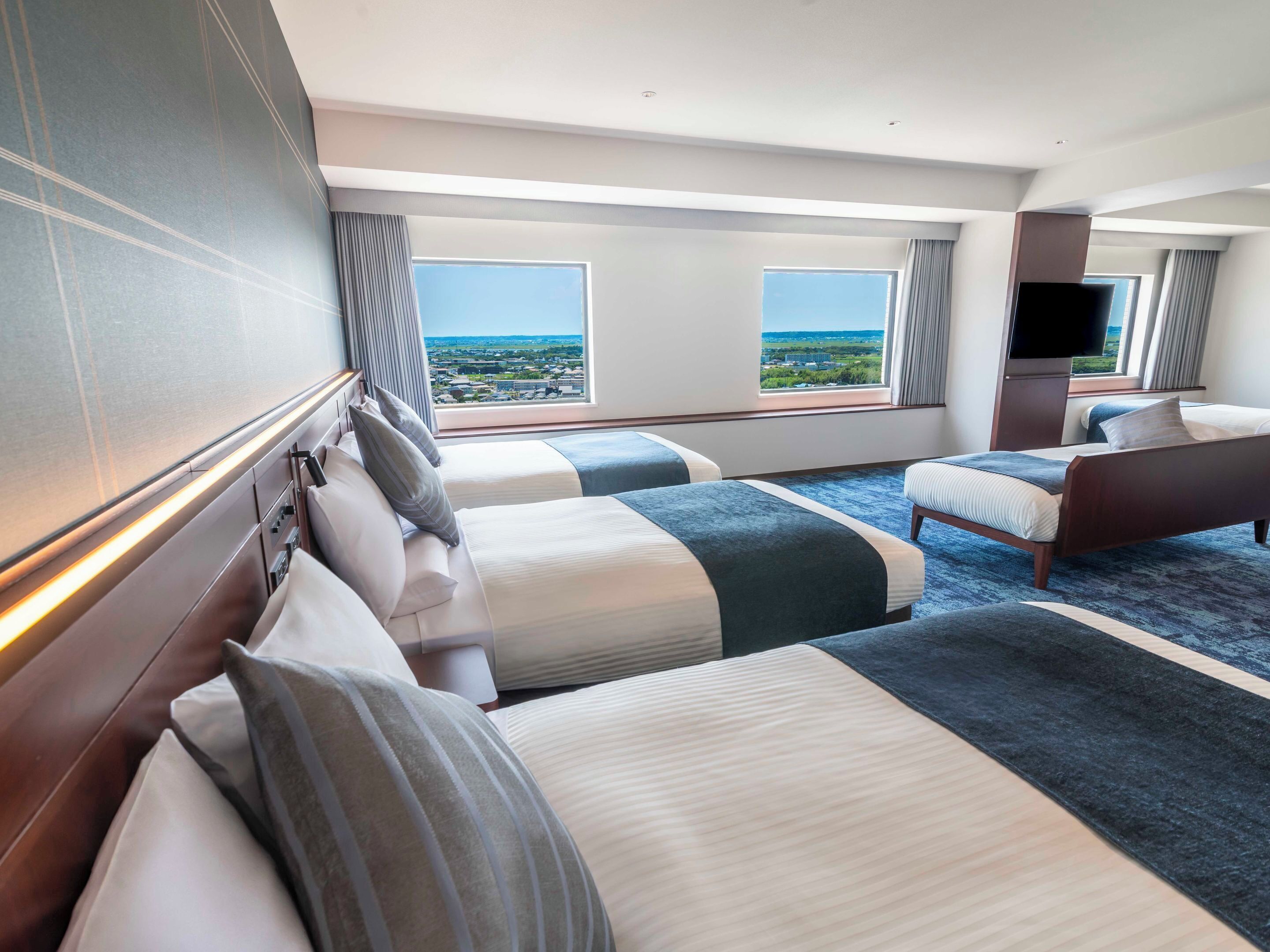 A hotel room with two beds, a flat-screen TV, and large windows offering a city view.