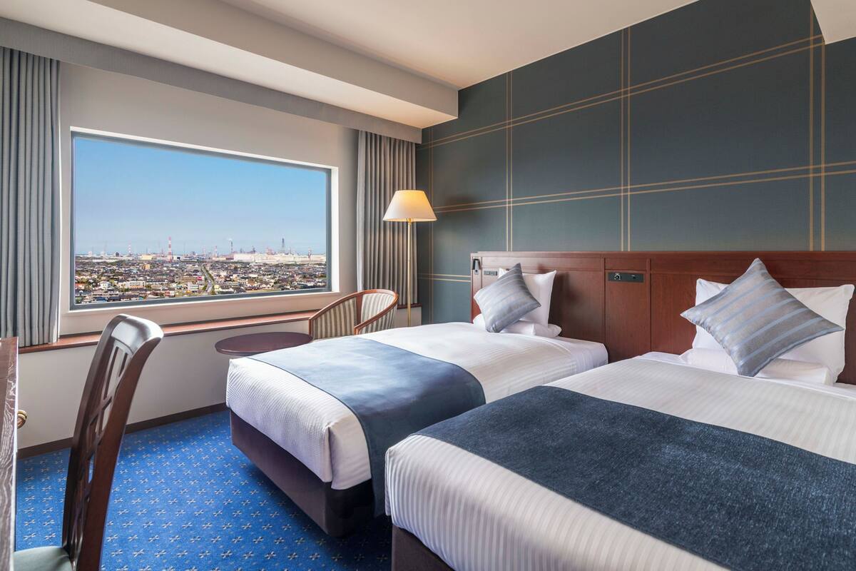 A hotel room with two beds, a view of a cityscape, a desk, and a chair.