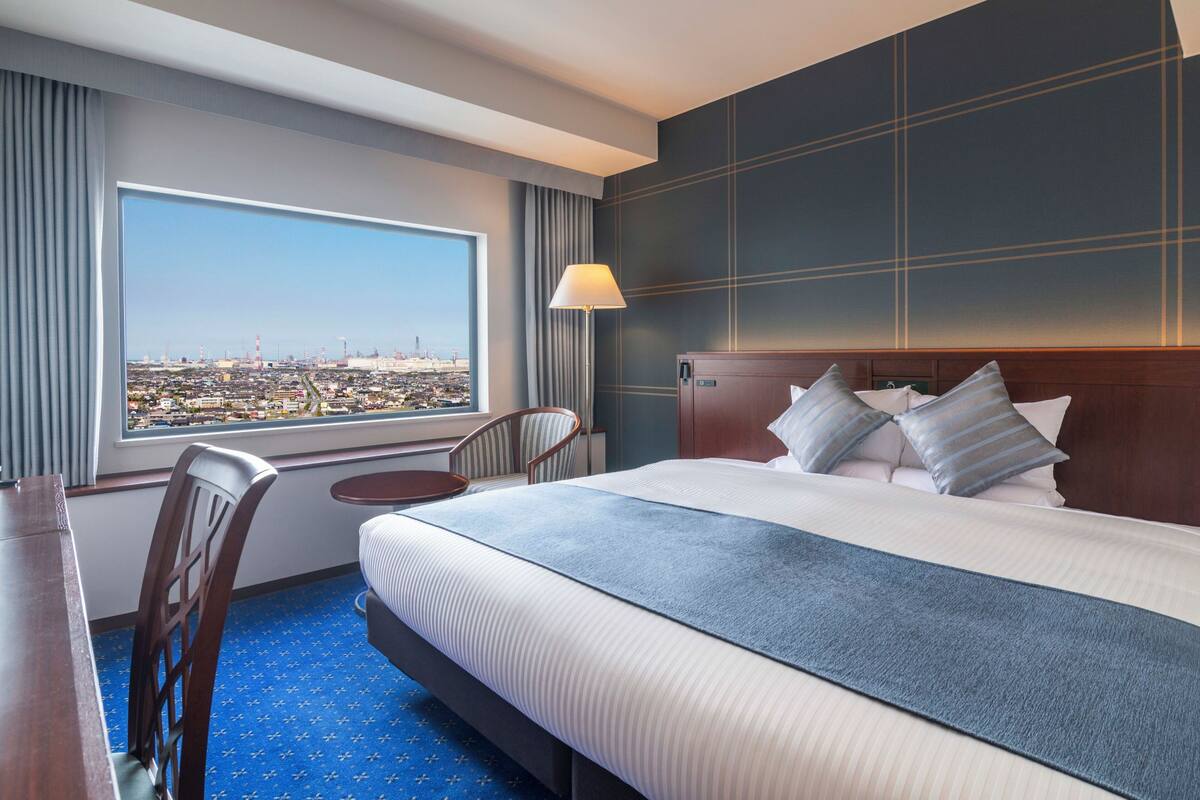 A hotel room with a large bed, a desk, a chair, a lamp, and a view of a cityscape through a large window.