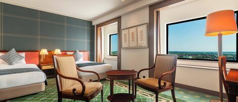 A hotel room with a bed, two armchairs, a small table, a floor lamp, and a large window with a city view.