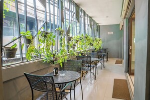A cafe with metal tables and chairs, potted plants, and a view through large windows.