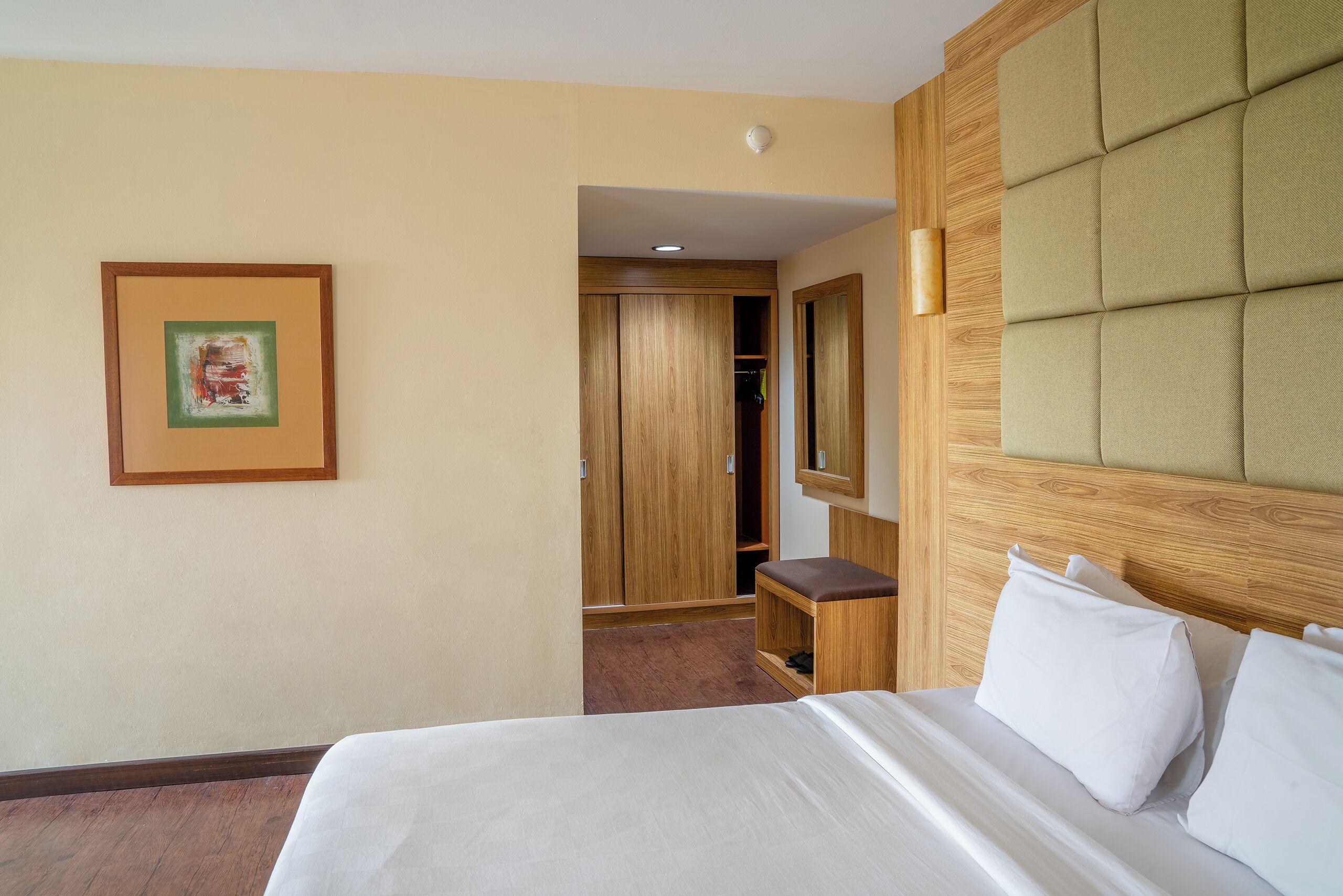 A hotel room with a bed, a wooden wardrobe, a mirror, and a framed picture on the wall.