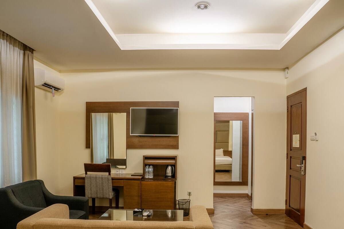 A hotel room with a sofa, a small table, a desk with a chair, a mirror, a television, and a door leading to another room.