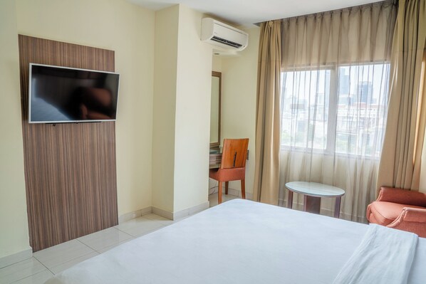 A hotel room with a bed, a TV mounted on a wooden panel, a chair, a small table, and a window with curtains. - Hotel Alia Cikini (Jakarta)