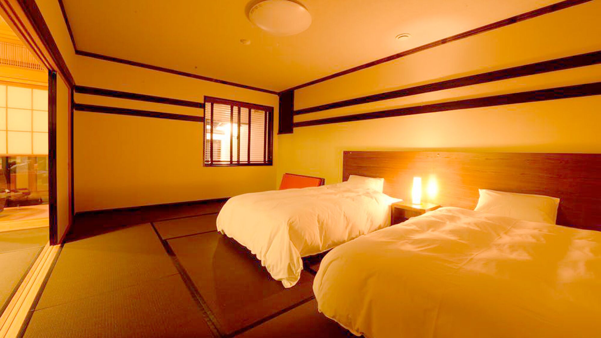 A hotel room with two beds, a wooden headboard, a window with blinds, and a tatami mat floor.