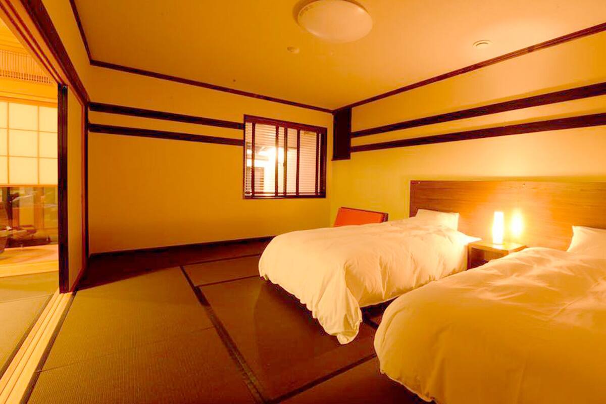 A hotel room with two beds, a wooden headboard, a window with blinds, and a tatami mat floor.