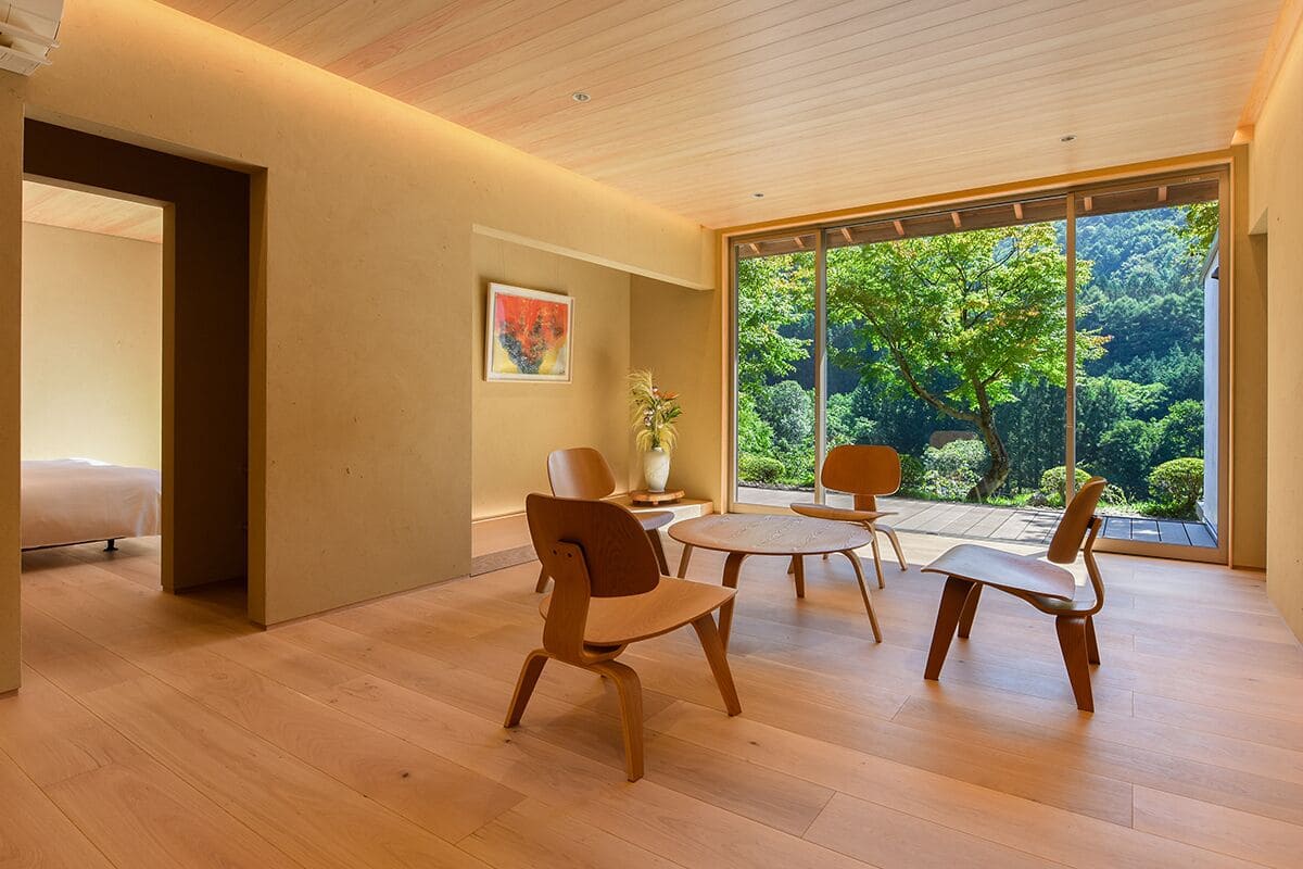 A modern interior with wooden flooring, a dining area with chairs, a bed, and a large window offering a view of greenery.