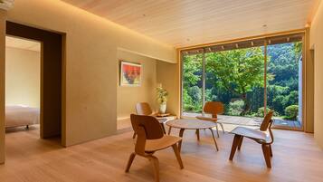 Interior moderno com soalho de madeira, área de refeições com cadeiras, uma cama e uma janela grande com vista para vegetação.