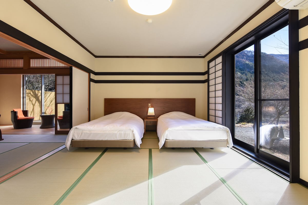 A traditional Japanese-style room with tatami flooring, two beds, a wooden headboard, and a large window offering a view of a mountainous landscape.