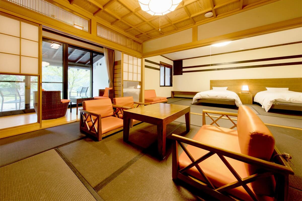 A traditional Japanese-style room with tatami flooring, a low wooden ceiling, and sliding shoji doors. There are two beds with white bedding, a wooden table, and several cushioned seating areas.