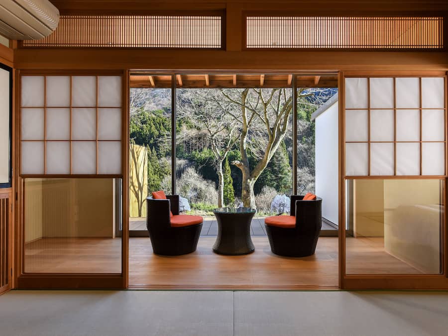 Una habitación japonesa tradicional con suelo de tatami, puerta corredera shoji y vistas a un área boscosa.