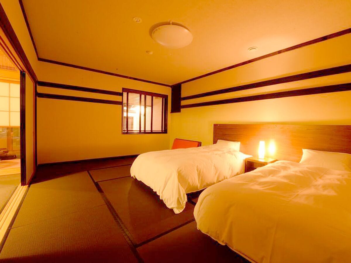 Una habitación con dos camas, una ventana con persianas, una mesita de noche con lámpara y suelo de tatami.