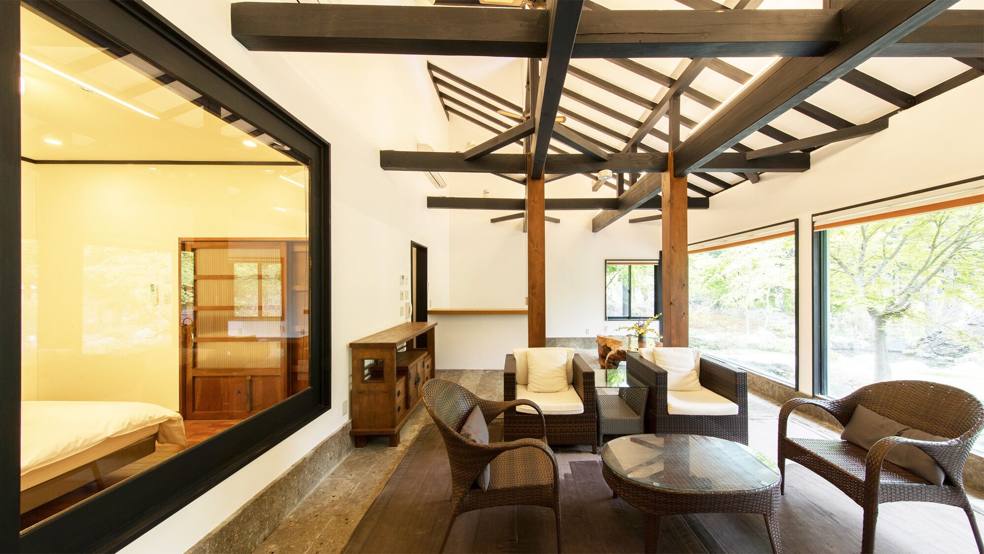 A modern interior with a glass wall, wooden ceiling beams, wicker furniture, and a view of greenery outside.