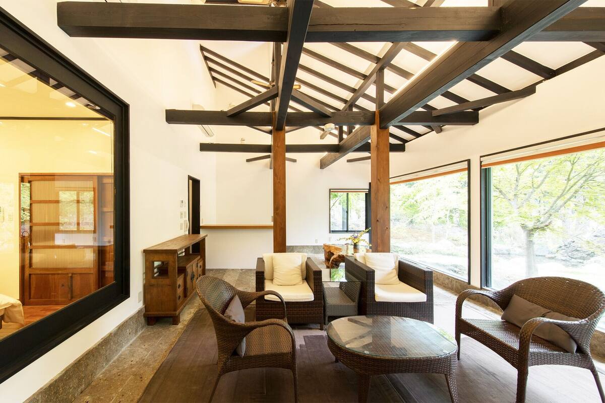 A modern interior with a glass wall, wooden ceiling beams, wicker furniture, and a view of greenery outside.