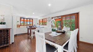 A modern kitchen with white cabinets, a wooden dining table, and a large window with a view of greenery. - Relax with your Best Friend in the heart of Tilba (Central Tilba)