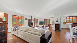 A spacious living room with a large sectional sofa, wooden flooring, and a kitchen area with white cabinets and a dining table. - Relax with your Best Friend in the heart of Tilba (Central Tilba)