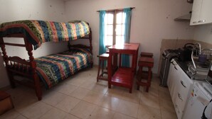 Een kleine, compacte kamer met een stapelbed, een houten tafel, twee krukjes, een raam met gordijnen en een kitchenette met een koelkast en fornuis.
