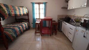 Een kleine kamer met een stapelbed, een tafeltje, een kitchenette met kookplaat en koelkast, en een raam met gordijnen.