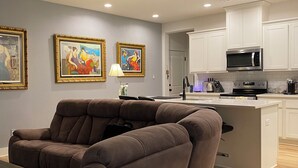 A modern living room with a brown sofa, a dining table, and a kitchen area with white cabinets and a microwave. - Charming townhome.  3-bdrm, 2.5 bath with pool. 10 minutes to Kyle Field. (College Station)