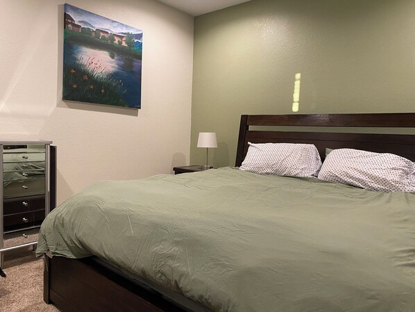 Een slaapkamer met een bed, nachtkastje, lamp, dressoir en een schilderij aan de muur.
