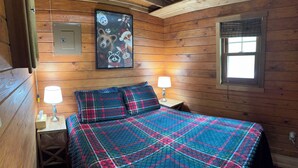 A wooden-paneled room with a bed, two bedside lamps, a framed picture on the wall, and a window with a blind. - Charming cabin in delightful Duquette with AC near miles of ATV trails. (Kerrick)
