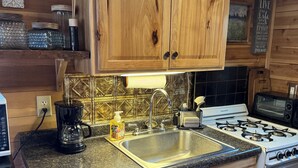A kitchen with wooden cabinets, a granite countertop, a stainless steel sink, a coffee maker, a toaster, a stove, and a microwave. - Charming cabin in delightful Duquette with AC near miles of ATV trails. (Kerrick)