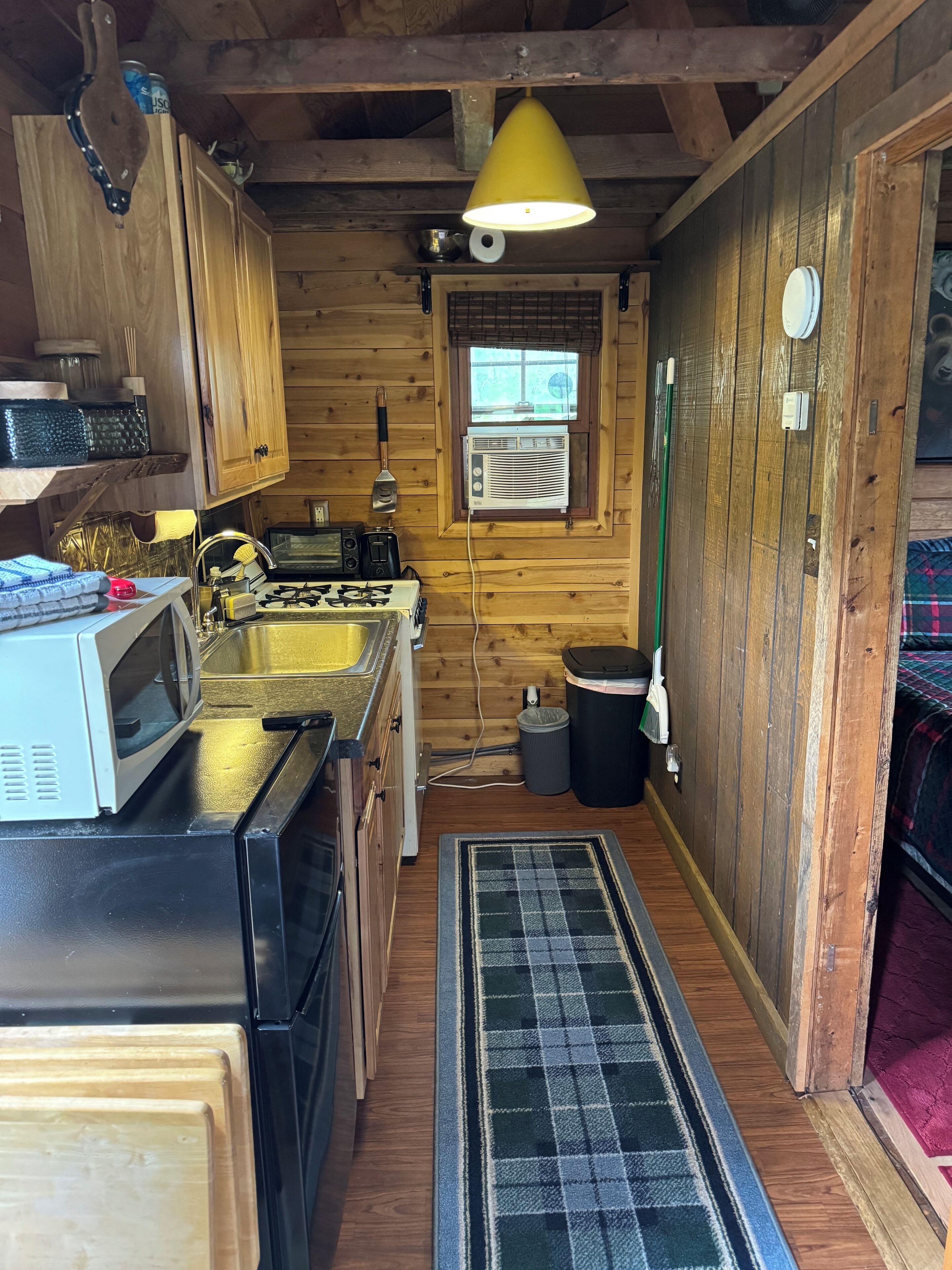 A compact kitchen with wooden walls, a microwave, stove, sink, and a window with blinds.