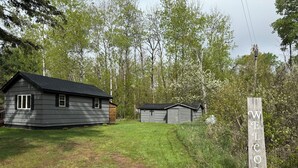 A rustic cabin with a wooden exterior and a welcoming 'WELCOME' sign, surrounded by lush greenery and trees. - Charming cabin in delightful Duquette with AC near miles of ATV trails. (Kerrick)